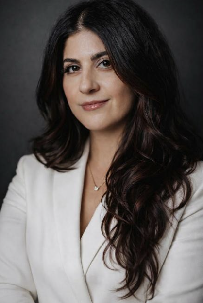 Mujer con cabello largo y ondulado, vistiendo un blazer blanco, posando sobre un fondo oscuro, mirando a la cámara con una expresión neutral.